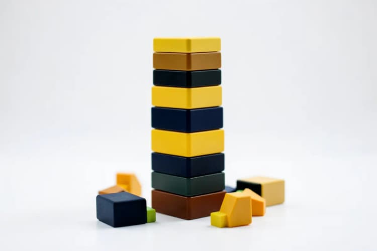 Uma pilha de blocos de construção coloridos formando uma torre instável, com alguns blocos caindo ao redor, em uma superfície branca.