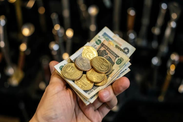 Uma mão segurando moedas e notas de dinheiro, com um fundo desfocado de ferramentas de vidraçaria.