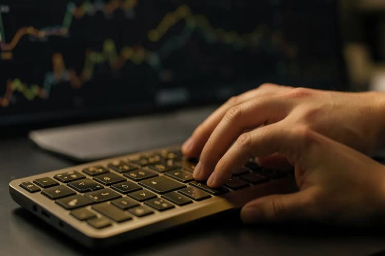 Mãos digitando em um teclado de computador com gráficos de análise de dados aparecendo em uma tela desfocada ao fundo.