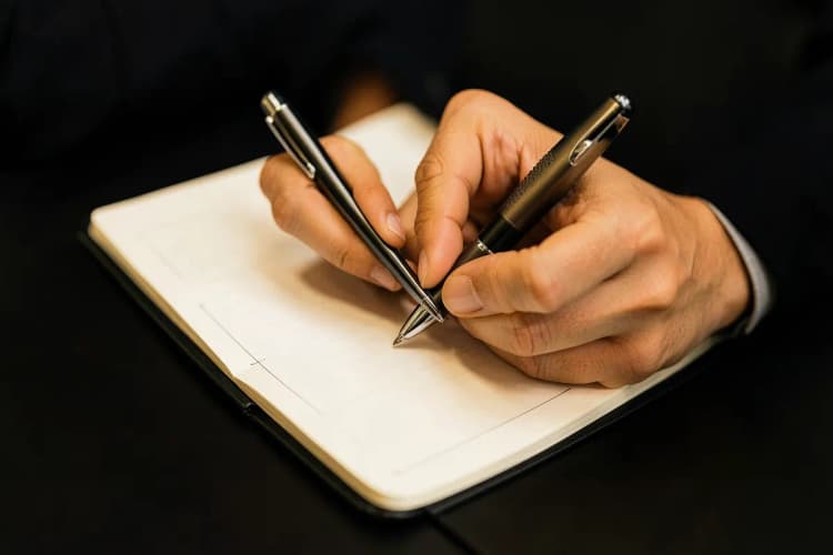 Uma pessoa escrevendo em um caderno com uma caneta, com uma xícara de café ao lado.