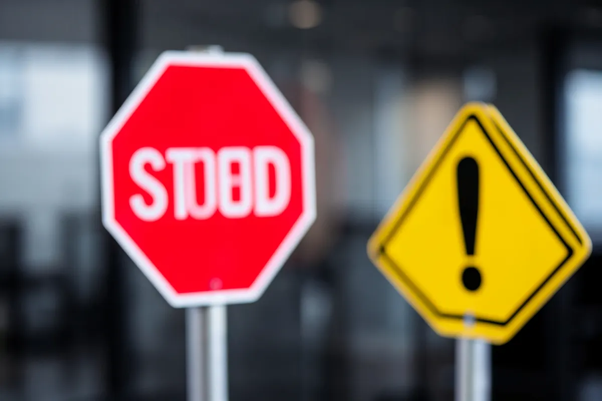 Uma placa de "Pare" vermelha e um sinal de "Cuidado" amarelo com um ponto de exclamação, sobre um fundo desfocado, simbolizando armadilhas.