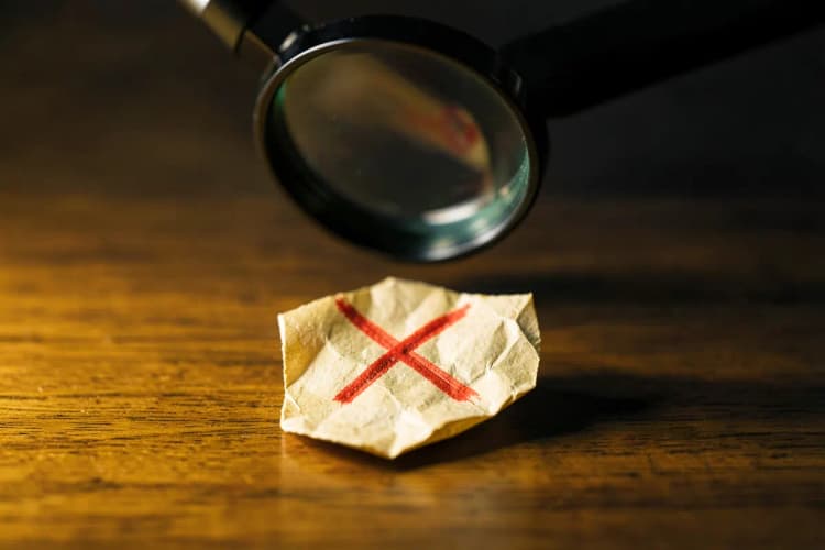 Uma lupa examinando um pequeno pedaço de papel amassado com um X vermelho desenhado, sobre uma superfície de madeira.